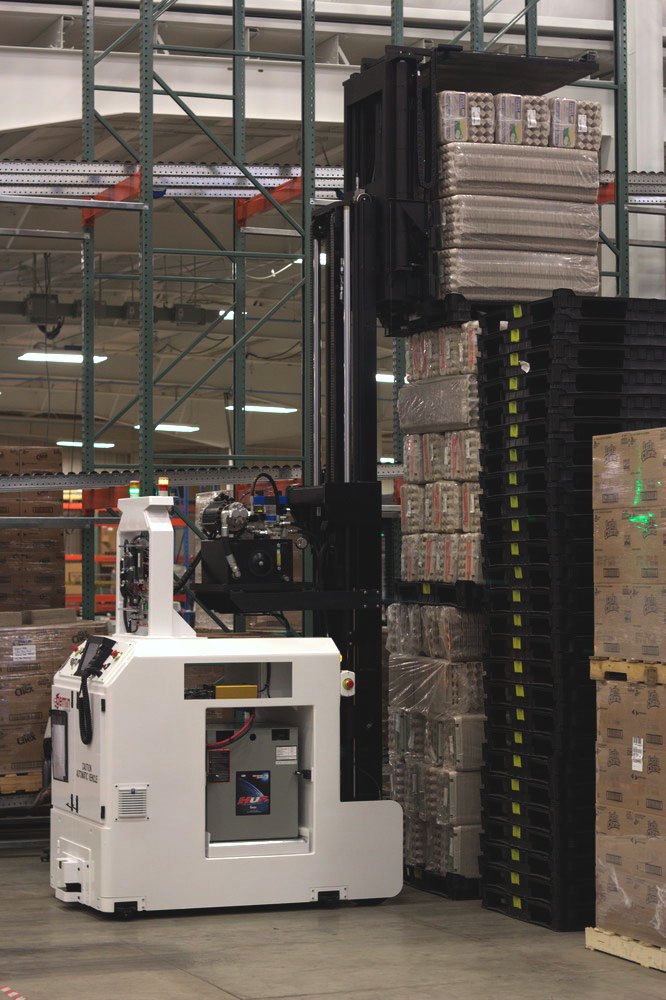 A warehouse automated guided vehicle (a low forklift robot) carrying a load down an aisle.