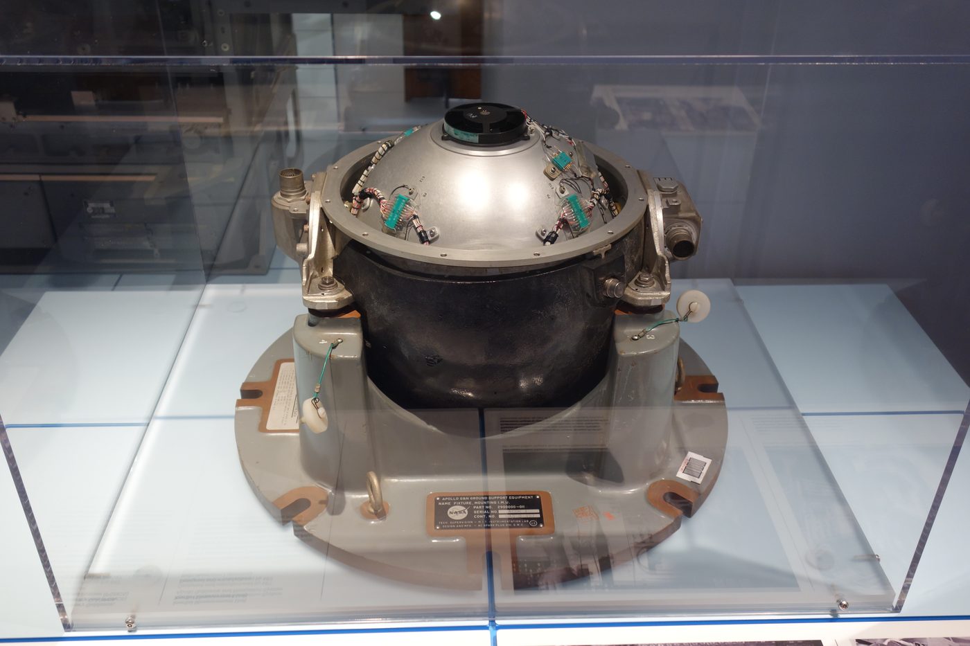 The spherical Apollo guidance-and-navigation inertial measurement unit on museum display, its polished case ringed with connectors and wiring.