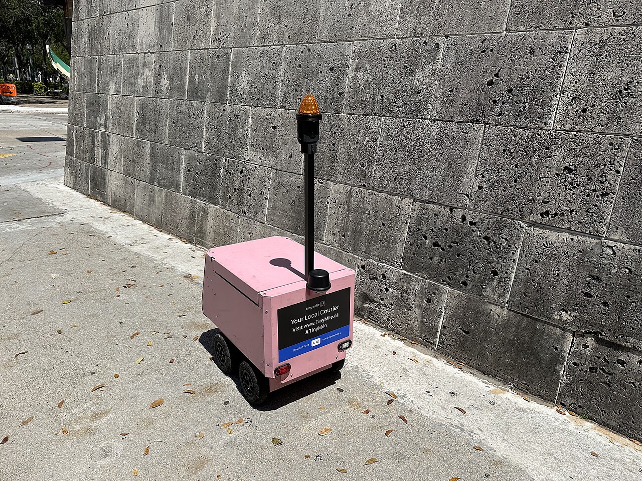 A small six-wheeled sidewalk delivery robot rolling along a pavement past buildings it has never mapped before.