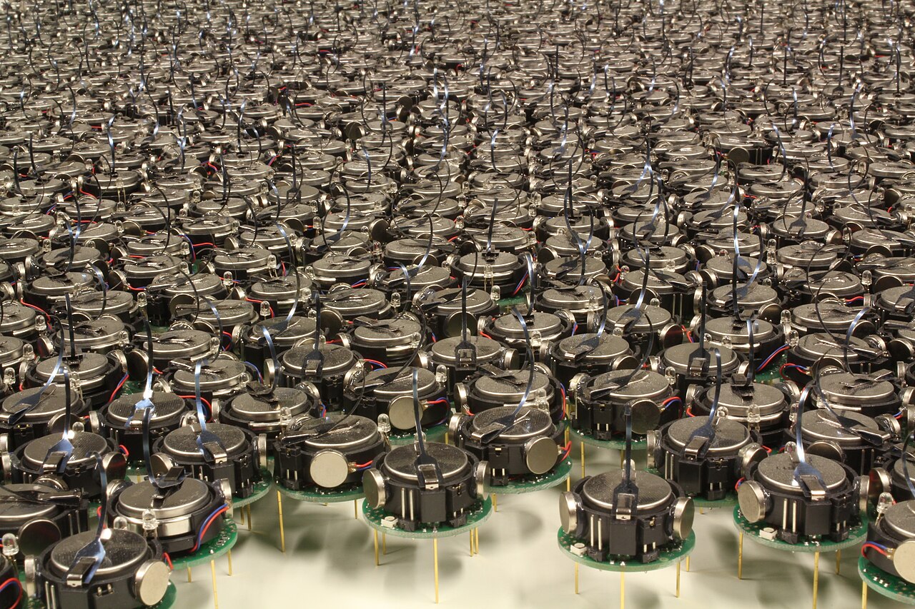 A dense swarm of tiny three-legged Kilobot robots clustered on a flat surface, each a small disc on vibrating legs.